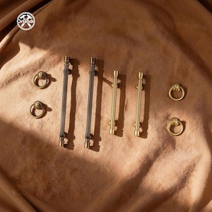 Op de afbeelding: Een collectie metalen kastbeslag bestaande uit twee donkerbronzen geribbelde staafgrepen, twee lichtere messing geribbelde staafgrepen en vier bijpassende ronde ringgrepen. Het Orlande-logo is zichtbaar in de linkerbovenhoek.