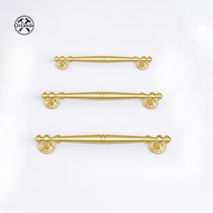 Puede incluir: Tres tiradores dorados para armarios con un diseño decorativo. Los tiradores están dispuestos verticalmente sobre un fondo blanco. La marca "Orlande" es visible en la esquina superior izquierda.