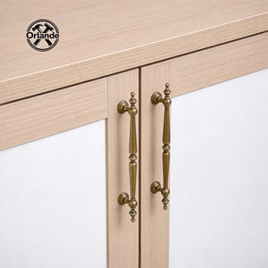 Può includere: Primo piano di un mobile con due maniglie in ottone antico. Le ante del mobile sono in legno chiaro con pannelli bianchi. La parte superiore del mobile è in legno chiaro. Il logo "Orlande" si trova nell'angolo in alto a sinistra.