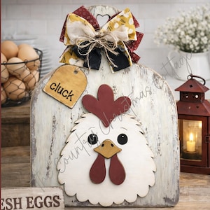 Puede incluir: Un letrero de madera blanca con forma de gallina y acabado desgastado, con cresta y barbillas rojas, pico amarillo y ojos negros. Un lazo decorativo corona la parte superior y una pequeña etiqueta de madera lleva la palabra cluck.