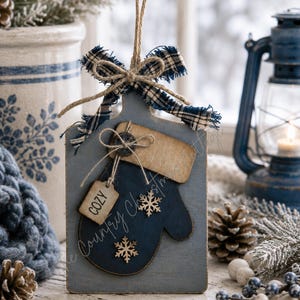 May include: A rectangular wooden sign featuring a dark blue mitten cutout, a plaid fabric bow, and two small wooden snowflake embellishments. A small tag labeled COZY is attached with twine. The sign includes text for a laser cut SVG file.