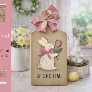 May include: A DIY wood kit featuring a bunny holding a tulip, with a pink bow and the words "SPRING TIME". The kit is ready to paint and includes a wood blank. A pink bow is at the top of the wood blank.