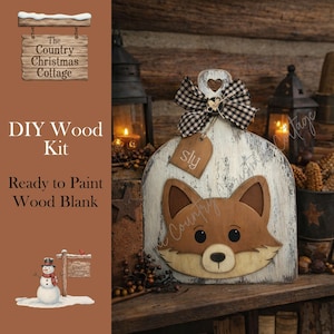 May include: A DIY wood kit featuring a fox design. The wooden blank is painted white with a distressed finish and a brown fox face. It includes a black and white checkered bow and a small tag that says "sly". The Country Christmas Cottage logo is visible.