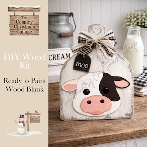 May include: A DIY wood kit featuring a ready-to-paint wood blank in the shape of a cow. The cow has a black and white face with pink ears and nose. A black tag says "moo". The kit is decorated with a bow.