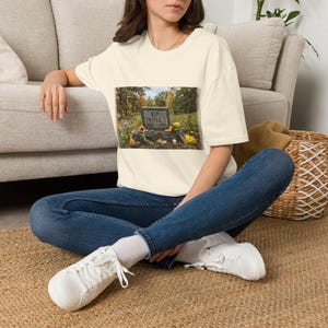 May include: An off-white short-sleeve t-shirt featuring a rectangular graphic print of a tombstone in a grassy field. The tombstone is inscribed with the text RIP TRIPLE T 1925-2026 and is surrounded by colourful flowers and autumn foliage.