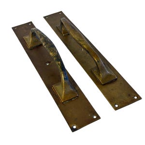 May include: Two rectangular brass door plates with attached arched handles. Each plate features a weathered, aged patina with visible dark spots and metallic texture. The plates have pre-drilled screw holes at the top and bottom for mounting.