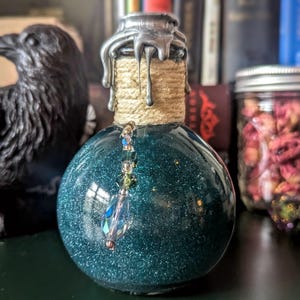 May include: A teal-colored, glittery glass bottle with a silver wax seal and a rope-wrapped neck. A decorative crystal pendant hangs from the bottle. The bottle is round and has a dark background with a raven statue and books.