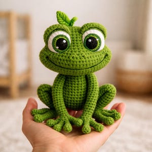 May include: A green crocheted frog with big eyes and a smiling expression. The frog has a small leaf on its head. This handmade item is a charming, whimsical creation, perfect as a decorative piece or a child's toy.