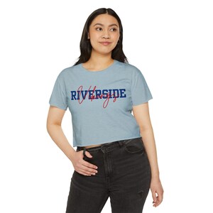 May include: Pale blue crop top featuring the text "Riverside" in navy blue and "life" in red script. A casual, short-sleeved top, ideal for everyday wear.