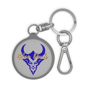 Op de afbeelding: Een ronde, grijze sleutelhanger met een blauw duivelsgezichtlogo en de woorden "Blue Devils" in gele letters. De sleutelhanger heeft een zilveren metalen ring en sluiting om aan sleutels of tassen te bevestigen.
