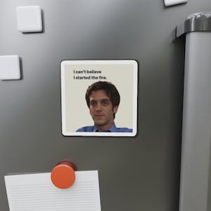 Puede incluir: Un imán cuadrado con un hombre de camisa azul y el texto "I can't believe I started the fire." El imán está adherido a una superficie gris.