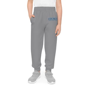 May include: Gray sweatpants with the word "VIKINGS" in blue script across the front. The pants have an elastic waistband and cuffs at the ankles. The person is wearing gray sneakers.
