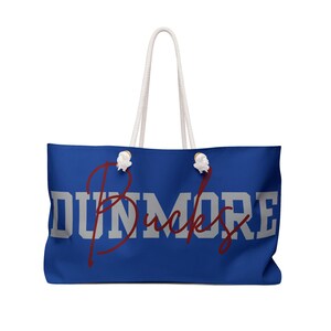 May include: A large blue tote bag with white rope handles. The bag features the word "DUNMORE" in large, gray block letters, with the word "Bucks" in a cursive, burgundy font overlaying it. The bag is designed for carrying items.