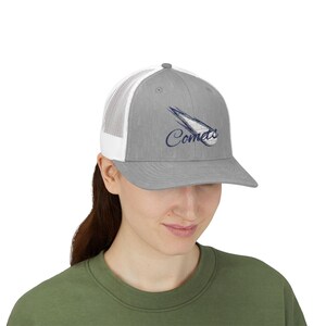 May include: A gray trucker hat with a white mesh back. The hat features the word "Comets" in a script font, along with a stylized comet graphic in navy blue. The hat is worn by a person.