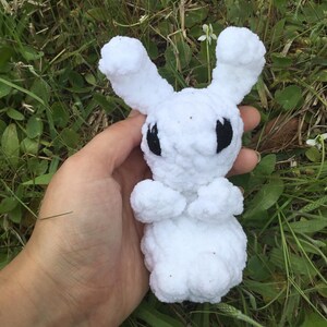 Può includere: Un coniglietto bianco fatto a mano all'uncinetto con occhi neri e lunghe orecchie. Il peluche è tenuto in mano su uno sfondo di erba verde e piccoli fiori bianchi. Il coniglietto è fatto di filato morbido e soffice.