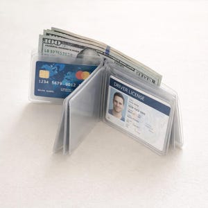 May include: A clear plastic wallet is open, displaying a credit card, a driver's license, and several one hundred dollar bills. The wallet has multiple clear pockets for organization.