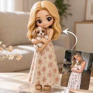 May include: A custom figurine featuring a blonde character with wavy hair wearing a cream-colored dress with a pink floral pattern, holding a small brown and white puppy. An arrow points to a photograph showing a person holding a similar puppy.