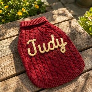 May include: A deep red cable-knit sweater for a pet featuring a ribbed collar and hem. The name Judy is embroidered in cream-colored yarn across the center of the garment in a cursive script font, adding a personalized touch to the textured knit.