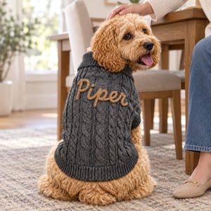 May include: A dark gray cable-knit dog sweater featuring a ribbed collar and hem. The back of the sweater is embroidered with the name Piper in a tan, cursive script font. The sweater is worn by a golden-colored dog with curly fur and a pink tongue.