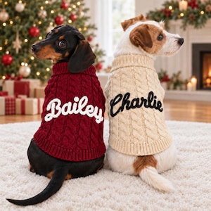 May include: Two dogs wearing cable-knit sweaters. The black and tan dog wears a deep red sweater with the white embroidered name Bailey. The white and brown dog wears a cream-colored sweater with the black embroidered name Charlie.