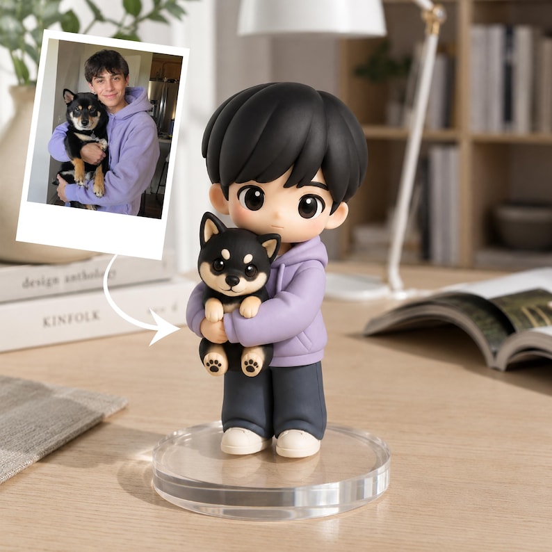 May include: A custom figurine of a person with black hair wearing a light purple hoodie and dark gray pants, holding a small black and tan Shiba Inu puppy. The figurine stands on a clear circular base and is modeled after the photo shown above it.