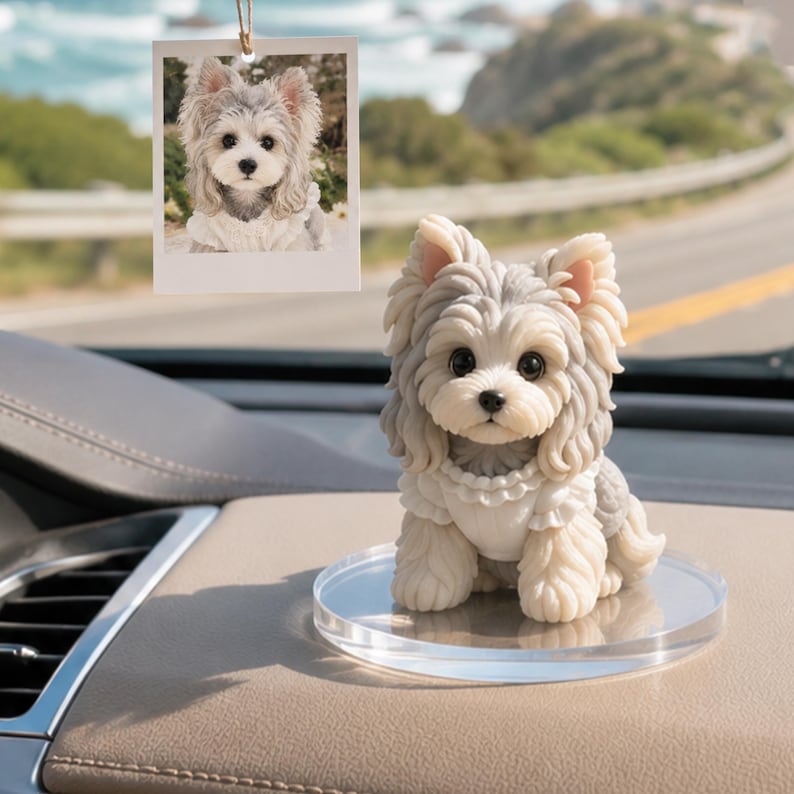 May include: A small, sculpted figurine of a Yorkshire Terrier with grey and cream fur, wearing a white ruffled collar, standing on a clear circular base. Above it hangs a square photo print of the same dog, suspended by a thin string.