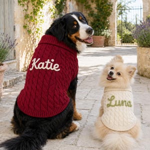 May include: A large Bernese Mountain Dog wearing a dark red cable knit sweater with the name Katie embroidered in white, and a small cream-colored Pomeranian wearing a matching cable knit sweater with the name Luna embroidered in green.