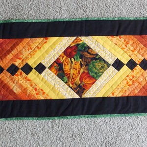 May include: A rectangular quilted table runner with a black border and a central design featuring a diamond pattern in shades of orange, yellow, and green. Black square accents are placed along the centre.