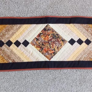 May include: A rectangular quilted table runner with a central diamond pattern. The runner has a black border with a rust-coloured trim. The centre design includes a floral print and various shades of brown, tan, and cream.