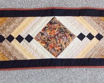 May include: A rectangular quilted table runner with a central diamond pattern. The runner has a black border with a rust-coloured trim. The centre design includes a floral print and various shades of brown, tan, and cream.