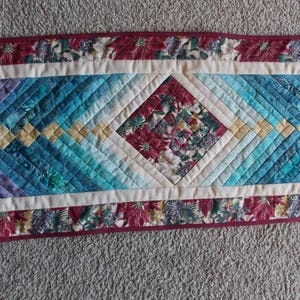 May include: A quilted table runner featuring a central diamond design in shades of blue, teal, and purple. The diamond is framed by a white border and a floral border in burgundy and cream. The runner is approximately 30 cm wide.