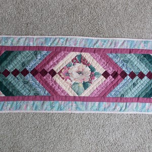 May include: A rectangular quilted table runner with a floral design. The centre features a diamond pattern with a large flower, surrounded by teal, pink, and burgundy fabrics. The edges have a floral print and a light blue border.