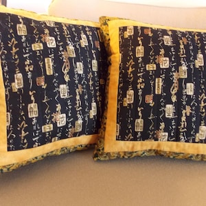 Könnte beinhalten: Zwei quadratische Kissen mit schwarzem Hintergrund und goldenen japanischen Schriftzeichen. Die Kissen haben einen goldenen Rand und liegen auf einem beigen Sofa. Dies sind dekorative Wohnaccessoires.