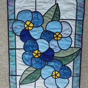 May include: A rectangular fabric wall hanging featuring a stained glass window design. The piece displays blue flowers with yellow centers and green leaves, outlined in black fabric against a white background with a blue and purple border.