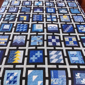 May include: A handmade quilt with a grid pattern of squares in various shades of blue, white, and black. Each square features a unique patchwork design, including geometric shapes and floral patterns. The quilt is displayed on a bed.