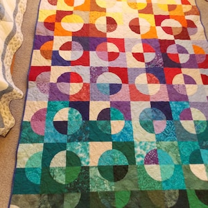 May include: A colorful handmade quilt with a geometric design. The quilt features squares with half-circle patterns in a variety of colors, including red, orange, purple, and teal. The quilt is likely made of cotton and is suitable for a bed or as a decorative throw.