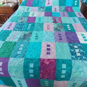 May include: A handmade quilt in shades of teal, purple, and green. The quilt features a patchwork design with rectangular blocks of varying patterns and textures. Some blocks have small, white square accents. The quilt is spread across a bed.