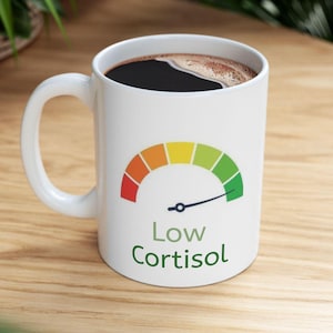 May include: White ceramic coffee mug filled with dark coffee. The mug features a colorful gauge design with the words "Low Cortisol" in green. The gauge ranges from red to green, indicating stress levels.
