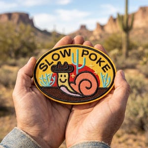 May include: An oval patch with the words "SLOW POKE" in yellow, featuring a cartoon snail wearing a cowboy hat. The snail is smiling, with a red and orange shell, and a cactus and sun graphic. The patch is held in cupped hands against a desert backdrop.