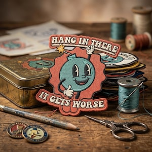 May include: Embroidered patch featuring a cartoon bomb character with a smiling face, thumbs up, and the text "HANG IN THERE" and "IT GETS WORSE". The patch is teal, orange, and white. Other patches, a pencil, and sewing supplies are in the background.