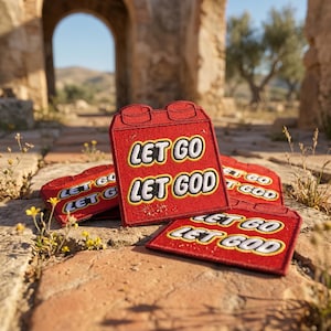 May include: Red embroidered patches in the shape of a building block, with the words "LET GO LET GOD" in white and yellow. The patches are arranged on a brick surface, with a blurred background of an archway and foliage.