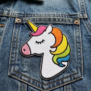 May include: Embroidered unicorn patch with a white body, pink nose, and a yellow horn. The unicorn's mane is a rainbow of colors, including pink, orange, yellow, green, and blue. The patch is sewn onto a denim jacket pocket.
