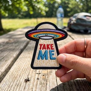 May include: Embroidered patch featuring a colorful UFO design with the text "TAKE ME" in red and blue. The UFO has a rainbow-colored top and a black border. The patch is held against a wooden surface.