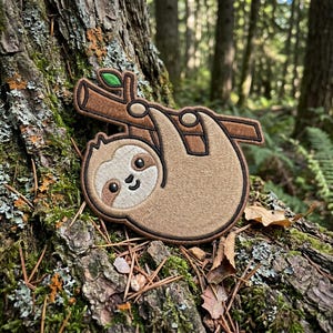 May include: Embroidered patch featuring a smiling sloth hanging from a brown branch with a green leaf. The sloth is light brown with a darker brown outline. The patch is set against a natural, wooded background.
