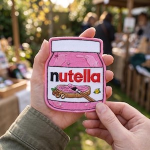 May include: Embroidered patch featuring a pink Nutella jar design. The patch includes the Nutella logo in red and white, along with an illustration of a slice of bread with Nutella, hazelnuts, and a yellow flower. The patch is held in a person's hands.