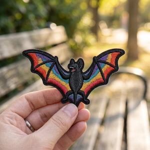May include: Embroidered bat patch with rainbow wings. The bat is black with open wings, showcasing vibrant rainbow colors. The patch is held in a hand against a blurred outdoor background.