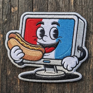 May include: Embroidered patch featuring a cartoon computer monitor with a smiling face, holding a hot dog. The monitor screen is colored red, white, and blue. The patch has a silver border and is set against a wooden background.