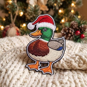 May include: An embroidered patch featuring a mallard duck wearing a red and white Santa hat. The duck has a green head, a brown chest, a white neck ring, and orange feet. The patch has a thick black border and detailed stitching throughout.