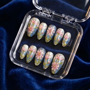May include: A set of ten oval-shaped press-on nails in a clear plastic case. Each nail features a hand-painted design of pink flowers with gold outlines on a light blue background. The nails are arranged on a dark blue velvet surface.