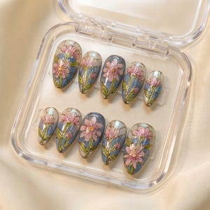 May include: A set of ten artificial nails in a clear plastic case. Each nail features a floral design with pink flowers, green leaves, and gold accents on a blue background. The nails are oval-shaped.